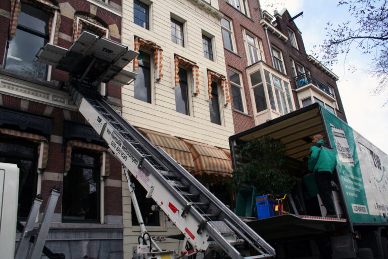 Een verhuislift vervoert meubels naar een bovenwoning in Utrecht met smalle gevel en verhuiswagen ervoor