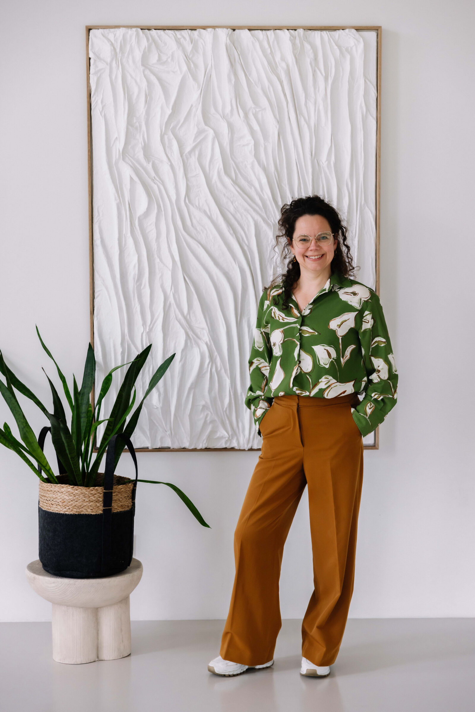 Els van Maren poseert lachend in haar studio naast een groene plant en een kunstwerk van wit textiel