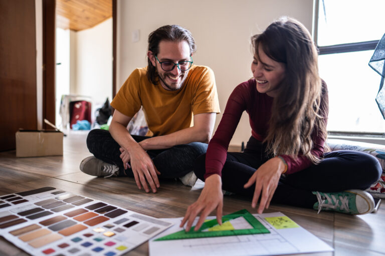 Stel zit lachend op de vloer van hun woning en bespreekt samen renovatieplannen met kleurstalen en bouwtekening