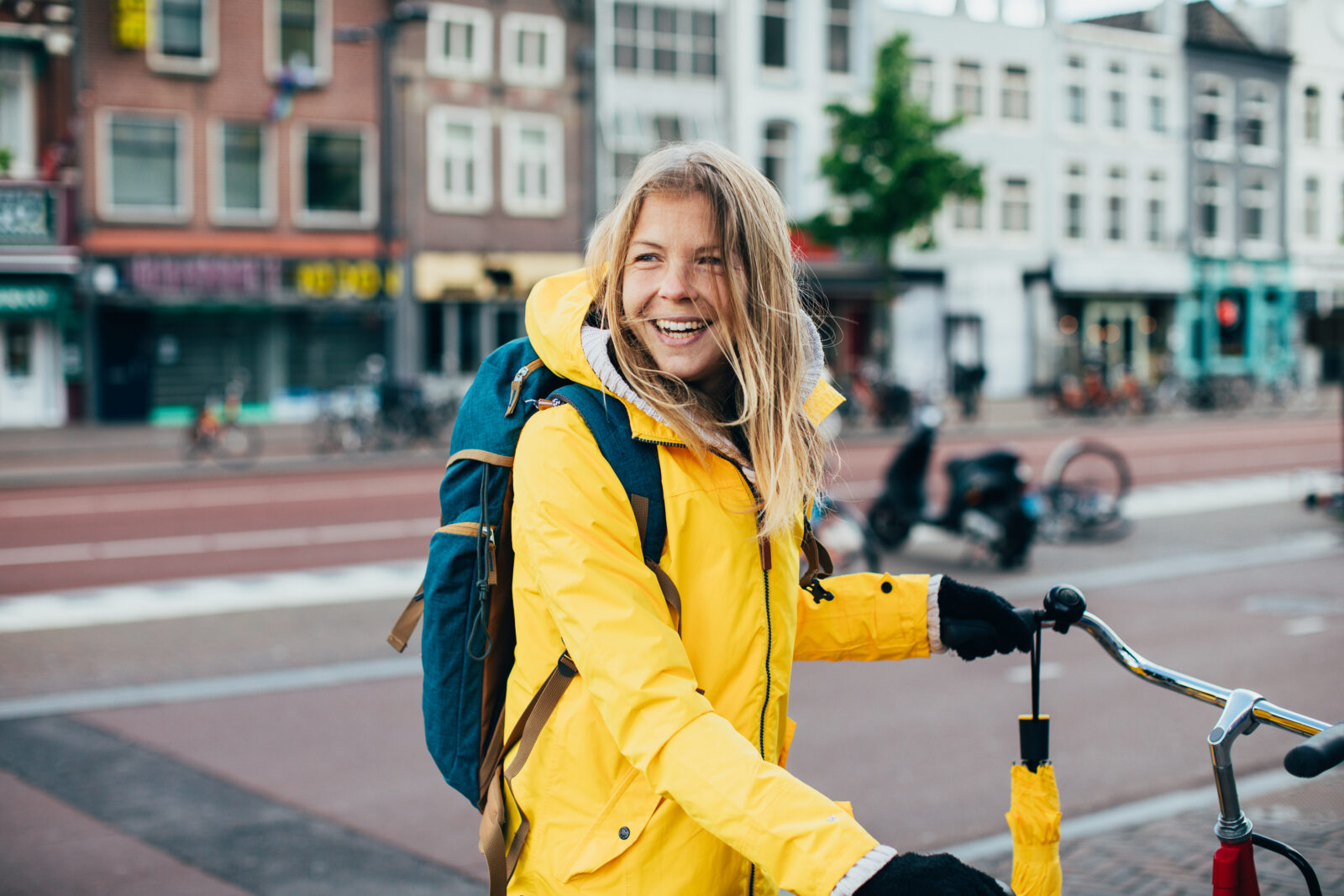 Lachende jonge vrouw op de fiets met gele jas en rugzak in een stadsomgeving