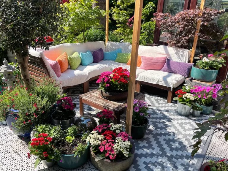 Kleurrijke loungeset met witte kussens en regenboogkleurige sierkussens, omringd door bloeiende planten in een zomerse tuin.