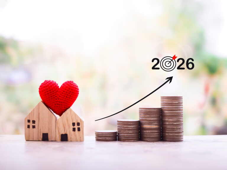 Houten huisjes met rood hart op muntstapels en pijl omhoog naar 2026 – symbool voor de keuze tussen kopen en huren.