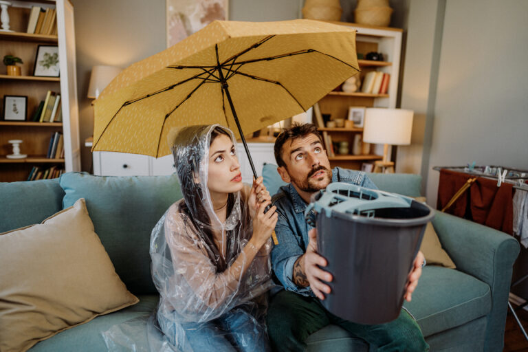 Stel onder een paraplu in de woonkamer terwijl water naar binnen lekt – symbool voor verbouwstress.