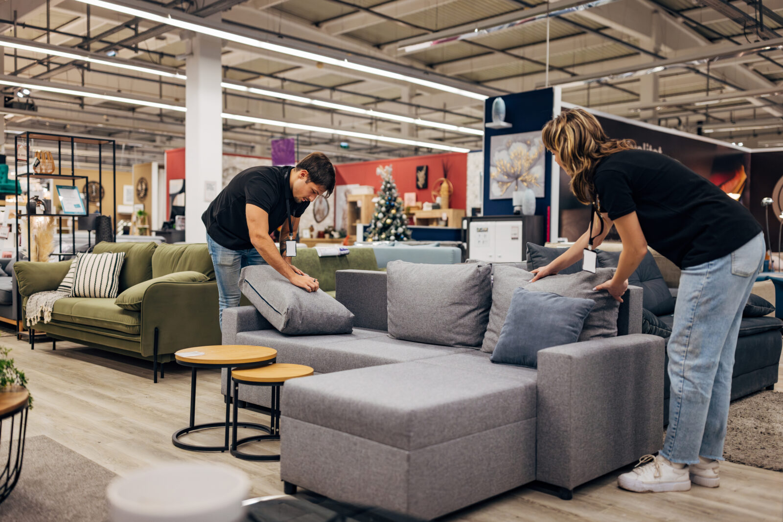 Twee mensen leggen kussens op een grijze bank in een meubelshowroom tijdens een comforttest.