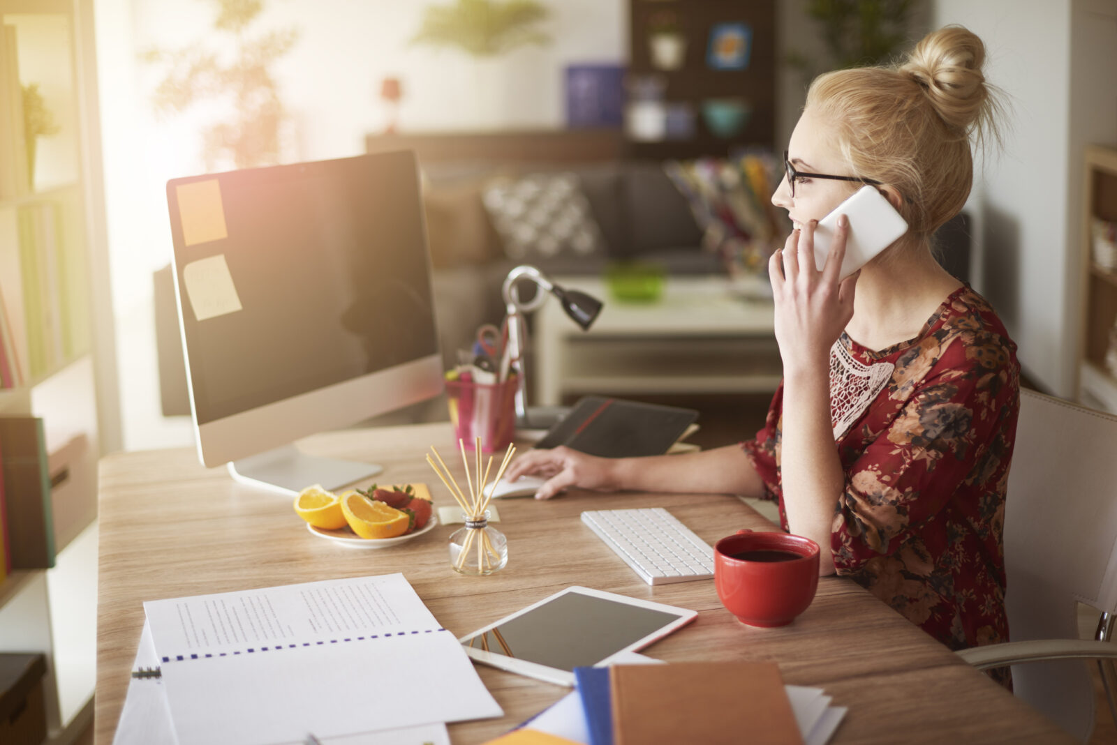 Werknemer in een thuiskantoor met fel zonlicht op het bureau, bezig aan de computer terwijl ze telefoneert.