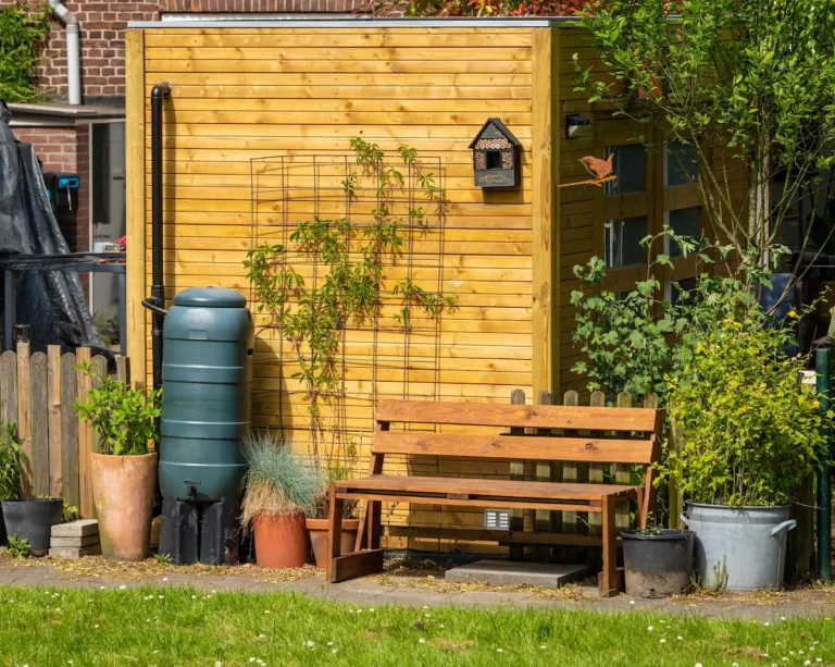Groene regenton naast houten schutting in een ingerichte tuin met bankje en planten.
