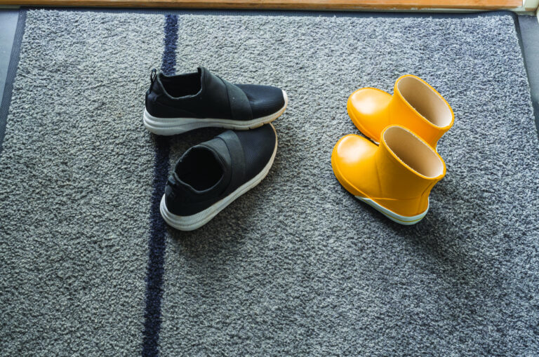 Schoenen en gele regenlaarsjes op grijze droogloopmat bij de voordeur