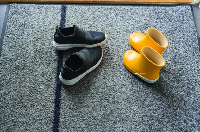 Schoenen en gele regenlaarsjes op grijze droogloopmat bij de voordeur