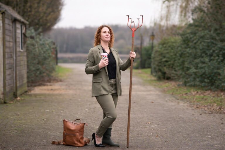 Aafke staat op een landelijk erf met een riek in haar hand en een kop koffie, omringd door groen en schuren op het platteland