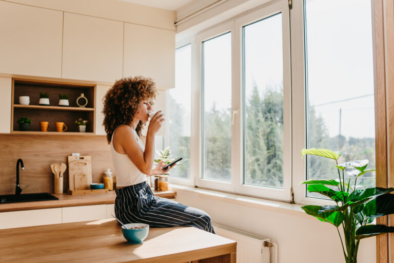 Vrouw zit op het aanrecht bij het raam met een kop koffie in een lichte moderne keuken